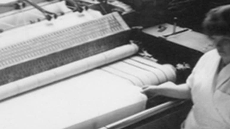 An old black and white photo of a woman working on a weaving machine.