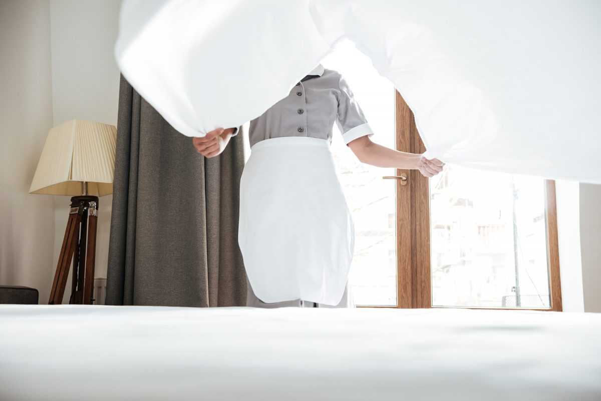 A maid in a white apron holding a sheet over a bed.