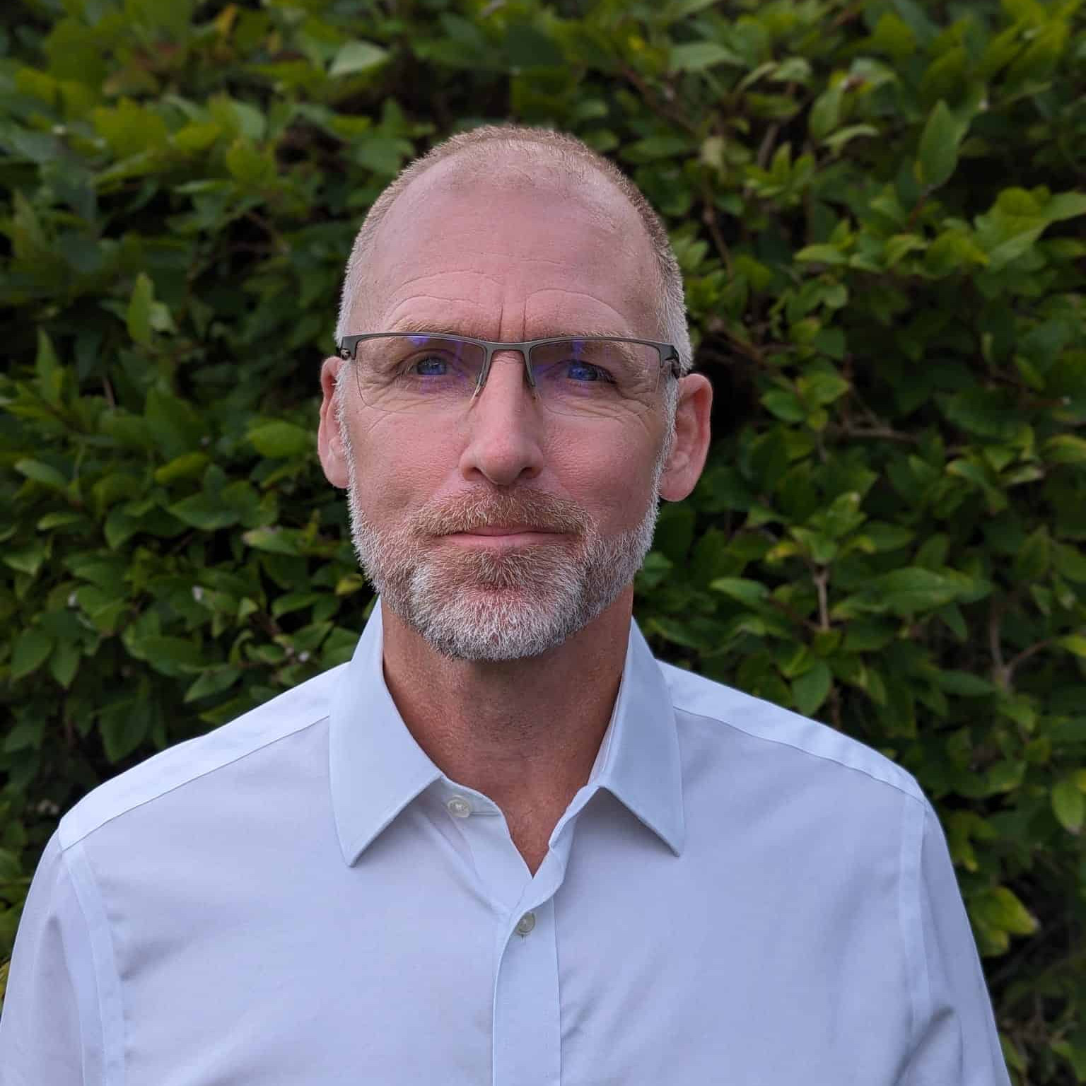 A man in a white shirt standing in front of bushes.