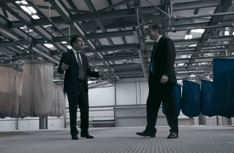 Richard & Mark Turvill are talking in an industrial setting, surrounded by large hanging textiles. The environment appears to be a warehouse or factory floor.